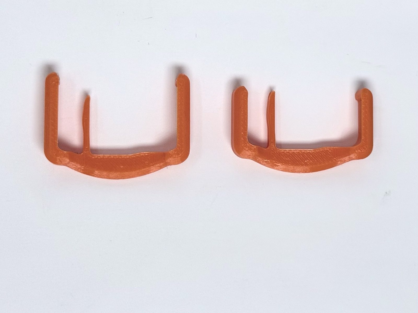 Two orange ResetClips side by side: the Gen 6 RC-106 on the left with longer arms, and the Gen 5 RC-105 on the right with shorter arms.